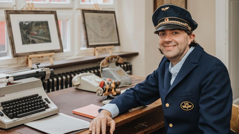 A man in a blue uniform with a cap is sitting at a desk with a typewriter and two telephones.