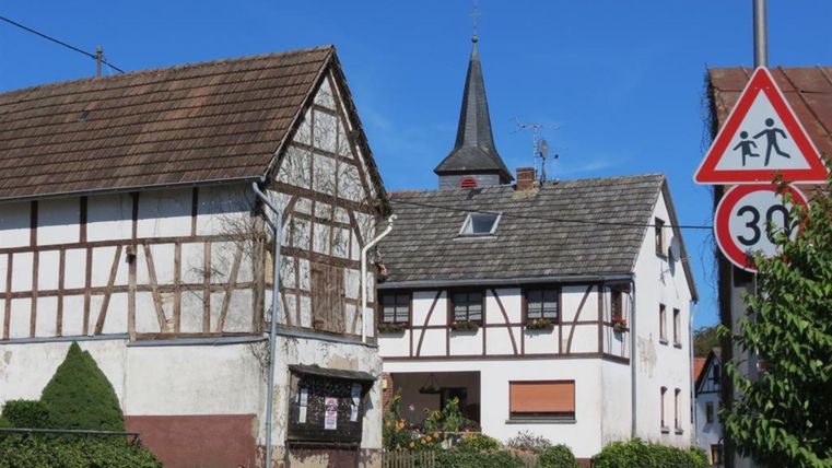 Ein malerisches Dorf mit Fachwerkhäusern und einem Kirchturm. Die Straße ist mit einem Geschwindigkeitsbegrenzungsschild versehen.