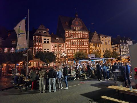 Ein belebter Marktplatz bei Nacht mit historischen, Fachwerkhäusern. Menschen stehen zusammen, während Stände Essen und Getränke anbieten.