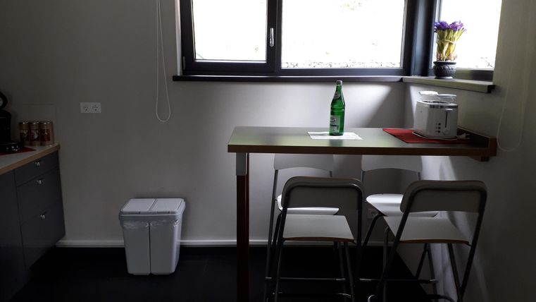 Eine moderne Küche mit einem kleinen Tisch und zwei Stühlen. Auf dem Tisch steht eine Wasserflasche und es gibt ein Fenster mit Blumen.