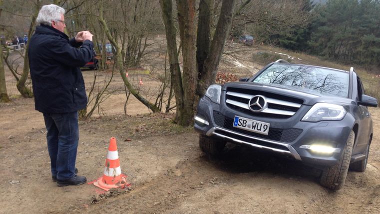 Ein Mann fotografiert ein geländetaugliches Mercedes-Fahrzeug auf einem unebenen Weg. Im Hintergrund sind Bäume und eine natürliche Umgebung sichtbar.