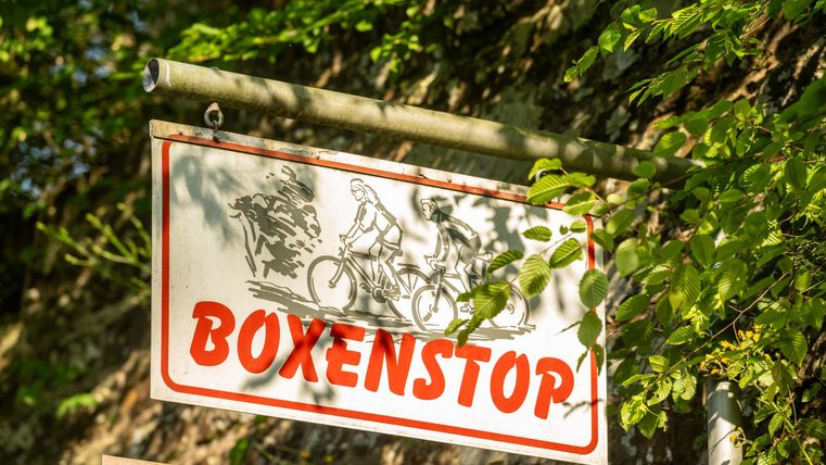 Ein Schild mit der Aufschrift "Boxenstop", das Radfahrer zeigt. Es ist umgeben von grünen Bäumen und Pflanzen.