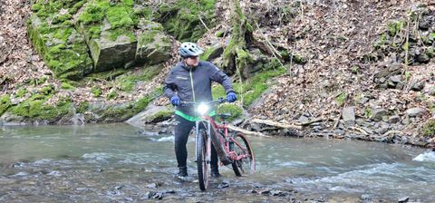 Ein Radfahrer überquert einen schmalen, fließenden Bach in einem bewaldeten Gebiet. Im Hintergrund sind moosbedeckte Felsen und trockene Blätter zu sehen.
