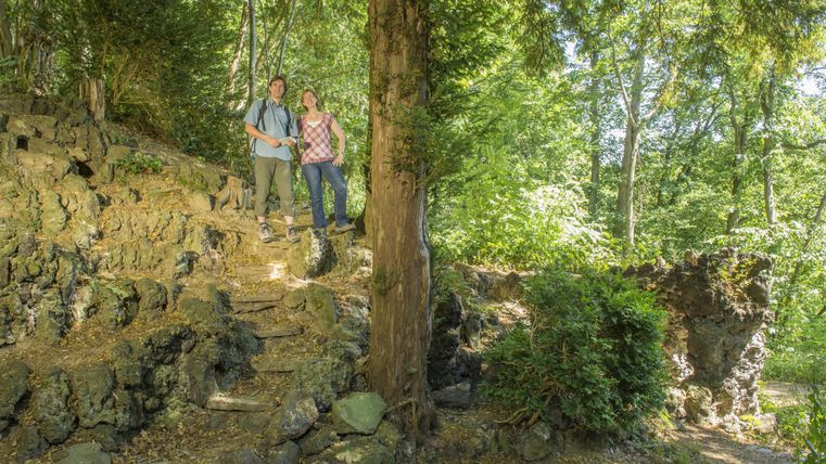 Ein Paar steht auf einer steinigen Treppe in einem sonnigen Wald. Umgeben von Bäumen und grünem Laub strahlt die Umgebung Ruhe und Naturverbundenheit aus.