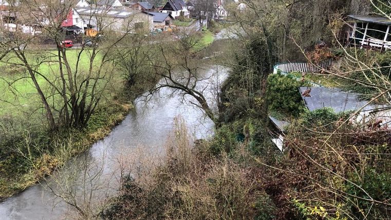 A quiet river flowing through a green landscape. In the background, houses and trees are visible.