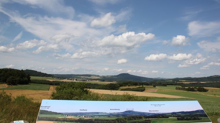 Aussicht von einer Plattform über eine ländliche Landschaft mit Feldern und Hügeln unter einem blauen Himmel mit Wolken.