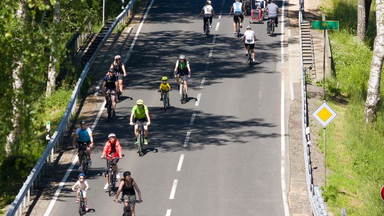 Eine belebte Fahrradstraße mit Radfahrern unterschiedlichen Alters. Die Sonne scheint und es gibt viel Grün um die Strecke herum.