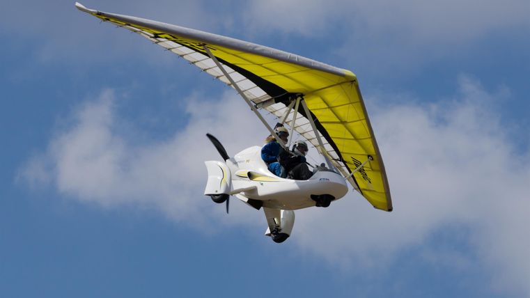 Ein leichtes Flugzeug mit gelbem Flügel, das in einem blauen Himmel fliegt. Der Pilot sitzt sichtbar im Cockpit.