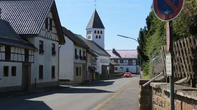 Ein ruhiger Weg in einem kleinen Dorf mit traditionellen Häusern. Im Hintergrund ist ein Kirchturm zu sehen.