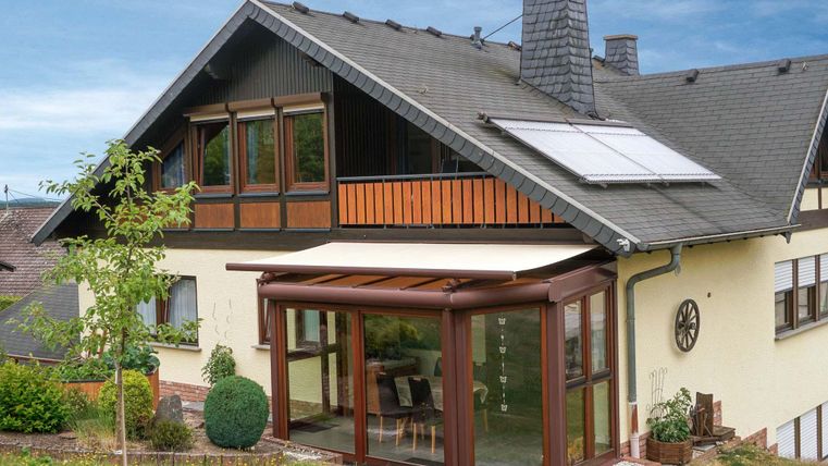 Ein modernes Haus mit einem Wintergarten und einer erneuerbaren Solaranlage auf dem Dach. Der Garten ist grün und gepflegt, was dem Gesamtbild eine einladende Atmosphäre verleiht.