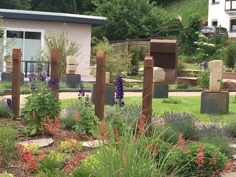 Un beau jardin avec différentes sculptures et des fleurs colorées. À l'arrière-plan, on aperçoit des bâtiments modernes et des plantes vertes.