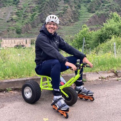 Een man zit op een klein, groen voertuig met grote banden. Hij draagt skateboard-uitrusting en lacht vriendelijk naar de camera.