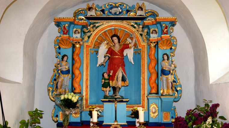 A colorful altar with the statue of an angel in red clothing. Around it are additional figures and decorative elements in blue and gold.