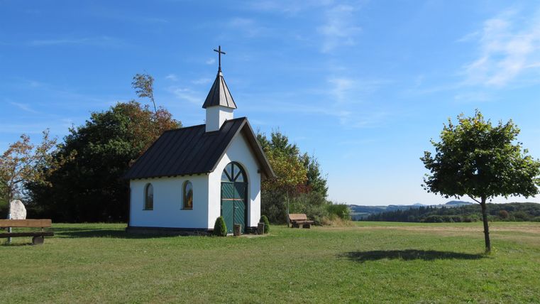Een kleine kerk staat op een groene weide onder een heldere blauwe lucht. Op de achtergrond zijn bomen en zachte heuvels zichtbaar.