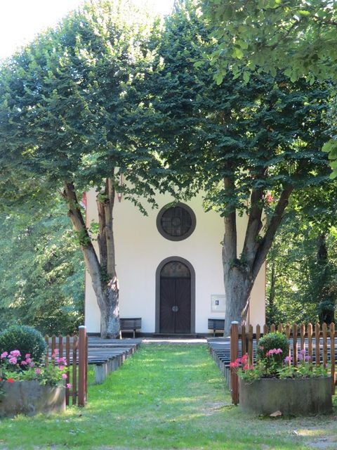 Eine kleine Kapelle mit einer Holztür, umgeben von großen Bäumen. Der Eingang ist von bunten Blumen und einer Holzumzäunung gesäumt.