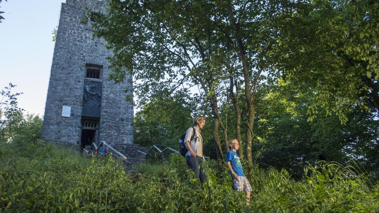 Eine Familie wandert an einem grünen Hang vorbei an einem alten Steinturm. Im Hintergrund sind Bäume und dichte Vegetation sichtbar.