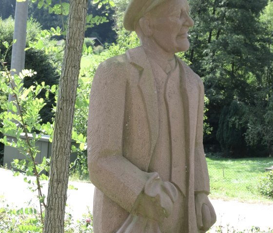 Steinfigur eines Mannes mit M&uuml;tze und Mantel, umgeben von B&auml;umen und Str&auml;uchern in einer l&auml;ndlichen Umgebung., &copy; Ti Adenau&copy;VGAdenau