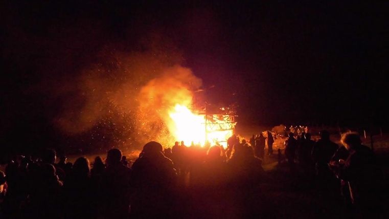 Ein großes Feuer brennt in der Nacht, umgeben von Menschen. Die Szenerie ist von orangefarbenem Licht und Funken erfüllt.