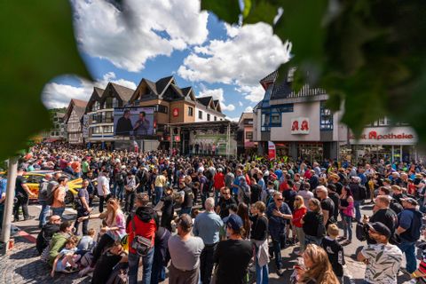 Eine große Menschenmenge versammelt sich auf einem Platz. Im Hintergrund sind Gebäude und eine Bühne zu sehen.