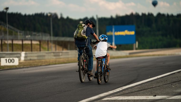 A father is riding bikes with his child. In the background, there is a natural landscape and a hot air balloon visible.