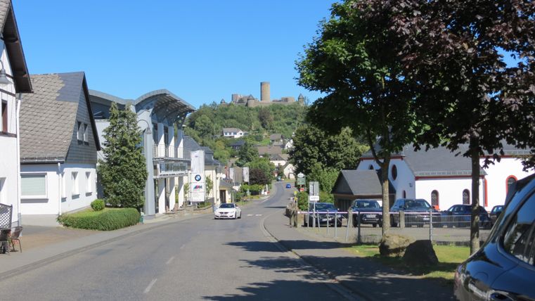Eine ruhige Straße mit weißen Häusern und Bäumen. Im Hintergrund ist eine Burgruine auf einem Hügel sichtbar.