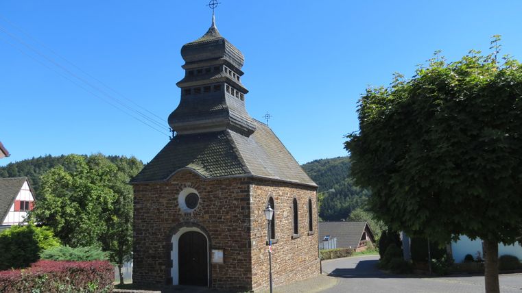 Eine kleine Steinkirche mit einem charakteristischen Dach steht an einer ruhigen Straße. Umgeben von grünem Lebensraum strahlt der Ort eine friedliche Atmosphäre aus.