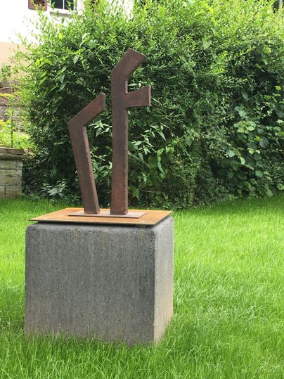 Une sculpture moderne se dresse sur un socle gris au milieu d'une herbe verte. En arrière-plan, des buissons sont visibles.