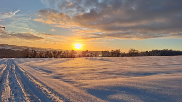 Een mooi winterlandschap met sneeuw en de zon die aan de horizon opkomt. De lucht is gedeeltelijk bewolkt en straalt in warme kleurtinten.