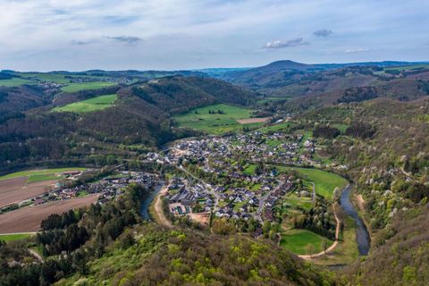 Eine malerische Landschaft mit einem kleinen Dorf, umgeben von sanften Hügeln und Wäldern. Ein Fluss schlängelt sich durch die grüne Wiese.
