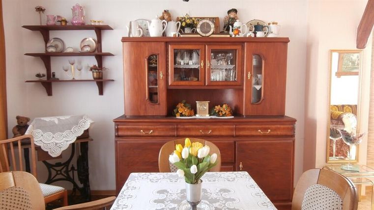 A cozy dining area with wooden furniture and a table decorated with a white tablecloth and a vase of flowers. In the background, there is a buffet with vintage dishes and decorative elements.