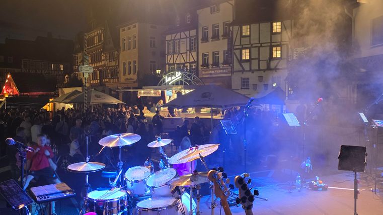 Eine Bühne mit Schlagzeug und Musikgeräten steht im Vordergrund. Im Hintergrund sind eine Menschenmenge und historische Gebäude bei Abenddämmerung zu sehen.