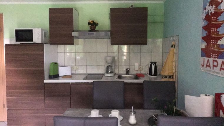 A modern kitchen with wooden furniture and light green walls. A table with chairs is in the foreground, and there are various kitchen appliances on the countertop.