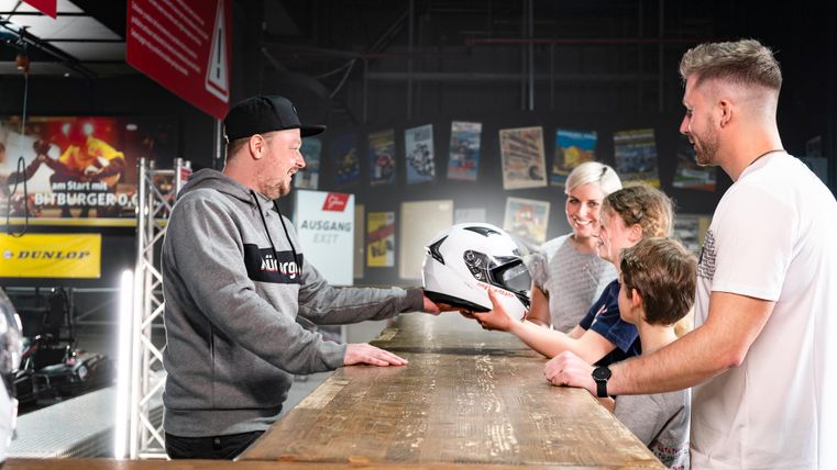 Eine Gruppe von Menschen steht an einem Holztisch und betrachtet einen Helm, der von einer Person übergeben wird. Die Atmosphäre wirkt freundlich und einladend.