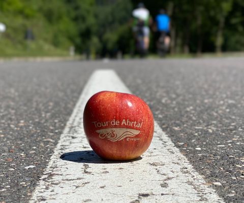 Ein roter Apfel mit der Aufschrift "Tour de Äpfel" liegt auf der Straße. Im Hintergrund sind Radfahrer zu sehen.