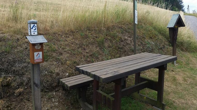 Ein einfacher Holztisch mit einer Bank steht in einer ländlichen Umgebung. Neben dem Tisch befinden sich Informationsschilder und eine kleine Hütte.