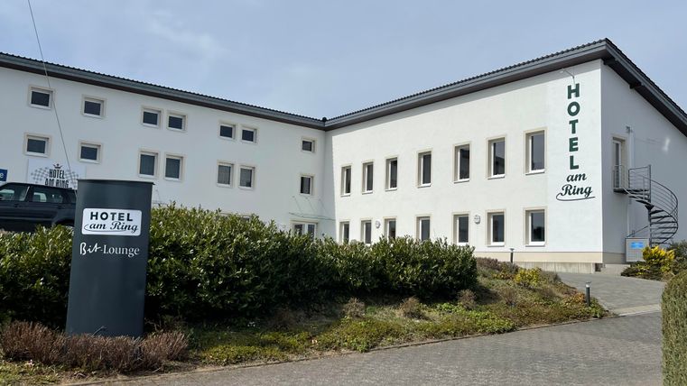 Een modern hotelgebouw met het opschrift "Hotel zur Ring". De gevel is licht en er is een uitnodigende omgeving met hagen.