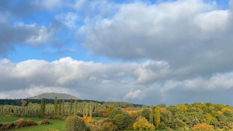 Un paysage pittoresque avec des prairies vertes et des arbres colorés en automne. Le ciel est nuageux avec quelques endroits lumineux.