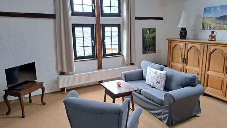 A bright living room with large windows and cozy furniture. Visible are a television, a table, and a spacious dresser.