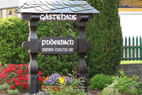 Een houten bord met de tekst "Gästehaus Puderbach" staat in een verzorgde tuin. Kleurige bloemen en een groene haag omlijsten de accommodatie.