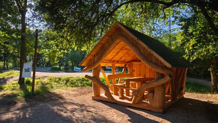 Eine gemütliche Holzhütte steht im Grünen, umgeben von Bäumen. Sie hat einen überdachten Sitzbereich und wirkt einladend.