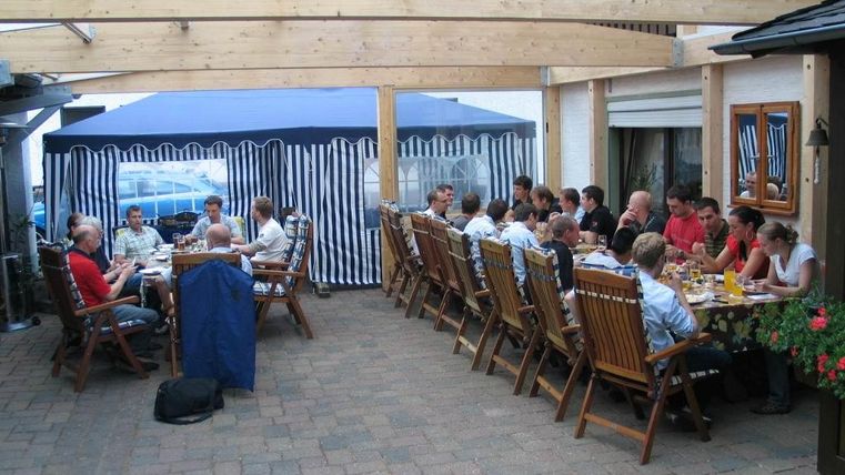 Een grote groep mensen zit aan lange tafels onder een overdekt gebied. Ze genieten van een gezamenlijke maaltijd in de buitenlucht.
