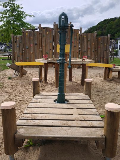 Ein Spielplatz mit einer Wasserpumpe in der Mitte. Im Hintergrund sind Kletterwände und ein Sandbereich zu sehen.