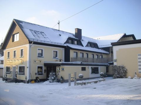 A cozy house in the snow, surrounded by a winter landscape. The sky is clear and the sun gently shines on the snowy surroundings.