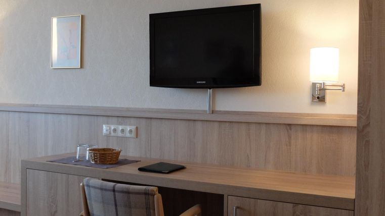 Een moderne kamer met een houten bureau, een tv aan de muur en een comfortabele stoel. Er is aangename verlichting en een kleine decoratie aan de muur.