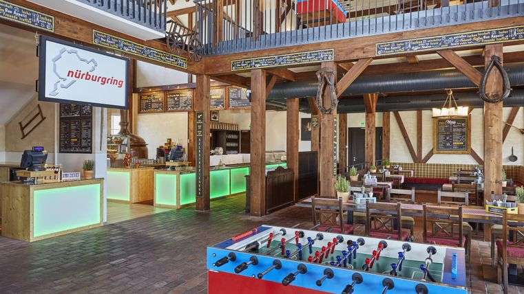 Un environnement de restaurant confortable avec un intérieur en bois rustique. Au premier plan, il y a un jeu de baby-foot et en arrière-plan, des tables pour manger.