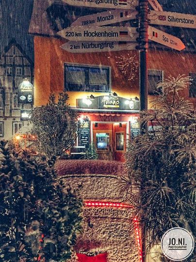 Ein gemütliches Restaurant mit einer einladenden Fassade und einem Schild mit Entfernungen zu verschiedenen Rennstrecken. Umgeben von vielen Pflanzen und festlicher Beleuchtung.