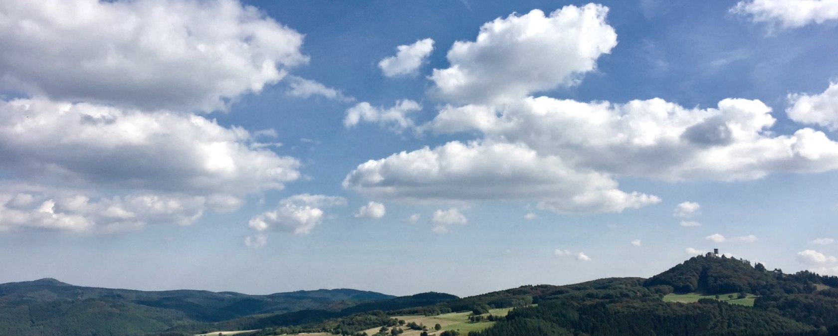 Weite Landschaft mit grünen Hügeln, Wäldern und Feldern. Auf einem Hügel ist die Burgruine Nürburg zu sehen. Der Himmel ist blau mit weißen Wolken., © VGVAdenau,SebastianSchulte