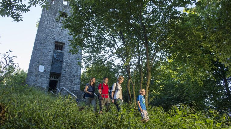 Eine Gruppe von Wanderern erkundet einen bewaldeten Hügel mit einem alten Turm im Hintergrund. Die Umgebung ist grün und sonnig, ideal für Outdoor-Aktivitäten.