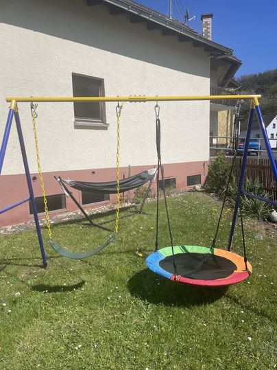 A playground with a colorful swing and a hammock is located in a garden. The background features a residential house and fresh greenery.