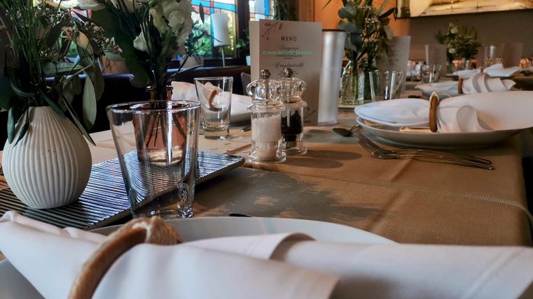 Ein eleganter gedeckter Tisch mit weißen Servietten und Blumendekoration. Im Hintergrund sind Glaswaren und Speisekarten sichtbar.